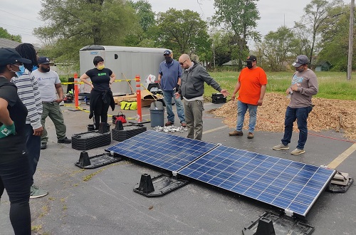 Group of people wearing masks standing outdoors around a large solar panel