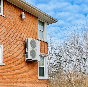 Side of a red brick multi-family building with a heat pump attached.