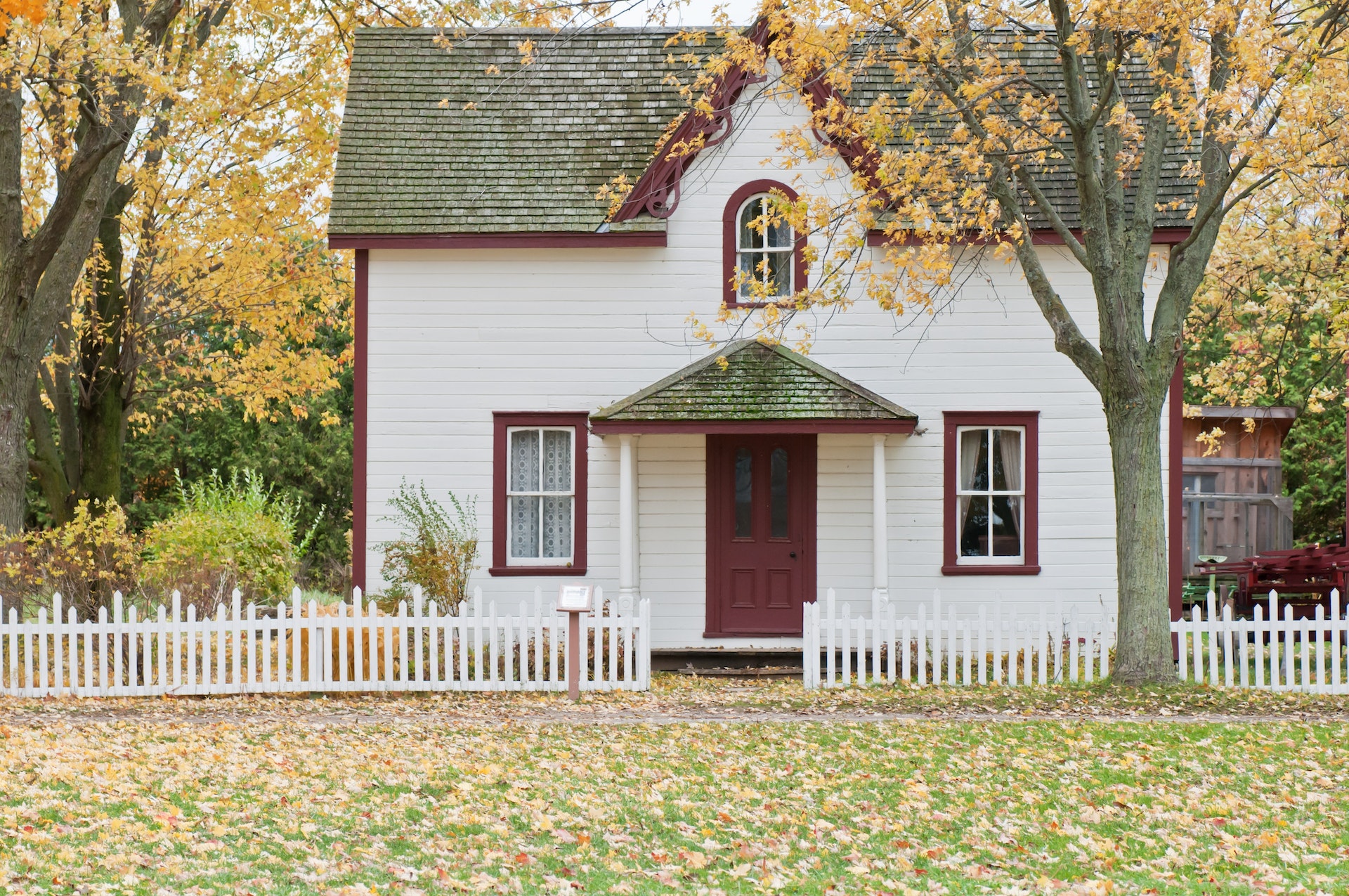 White and red wooden house with fence and trees with leaves changing color and falling.
