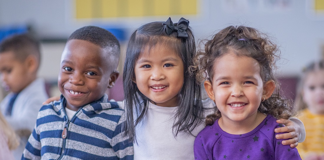 A multi-ethnic group of daycare children sit close together on the floor, with their legs crossed and their arms around one another, as they pose for a portrait. They are smiling and enjoying their time together.